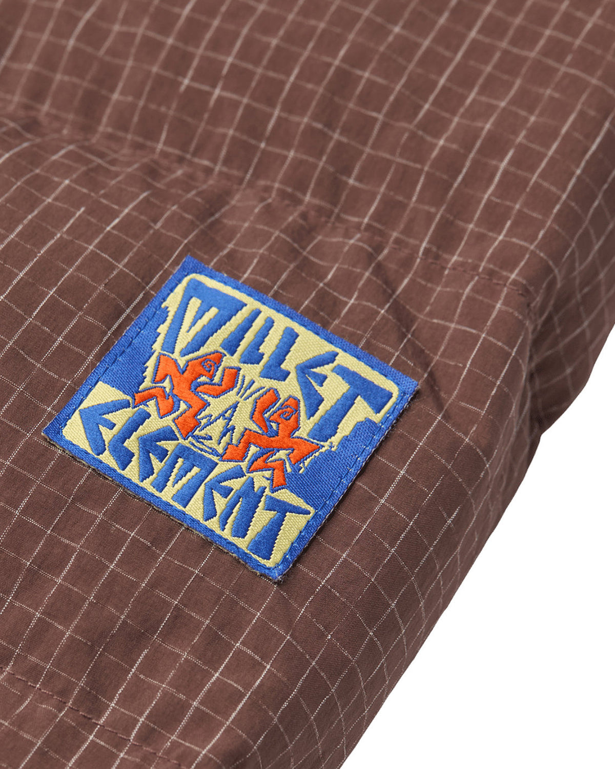 Vestes Element Mxe Puff Down J (N353) Truffle Mxe, gros plan sur un écusson carré bleu et jaune brodé du logo Millet Element, cousu sur le tissu quadrillé marron